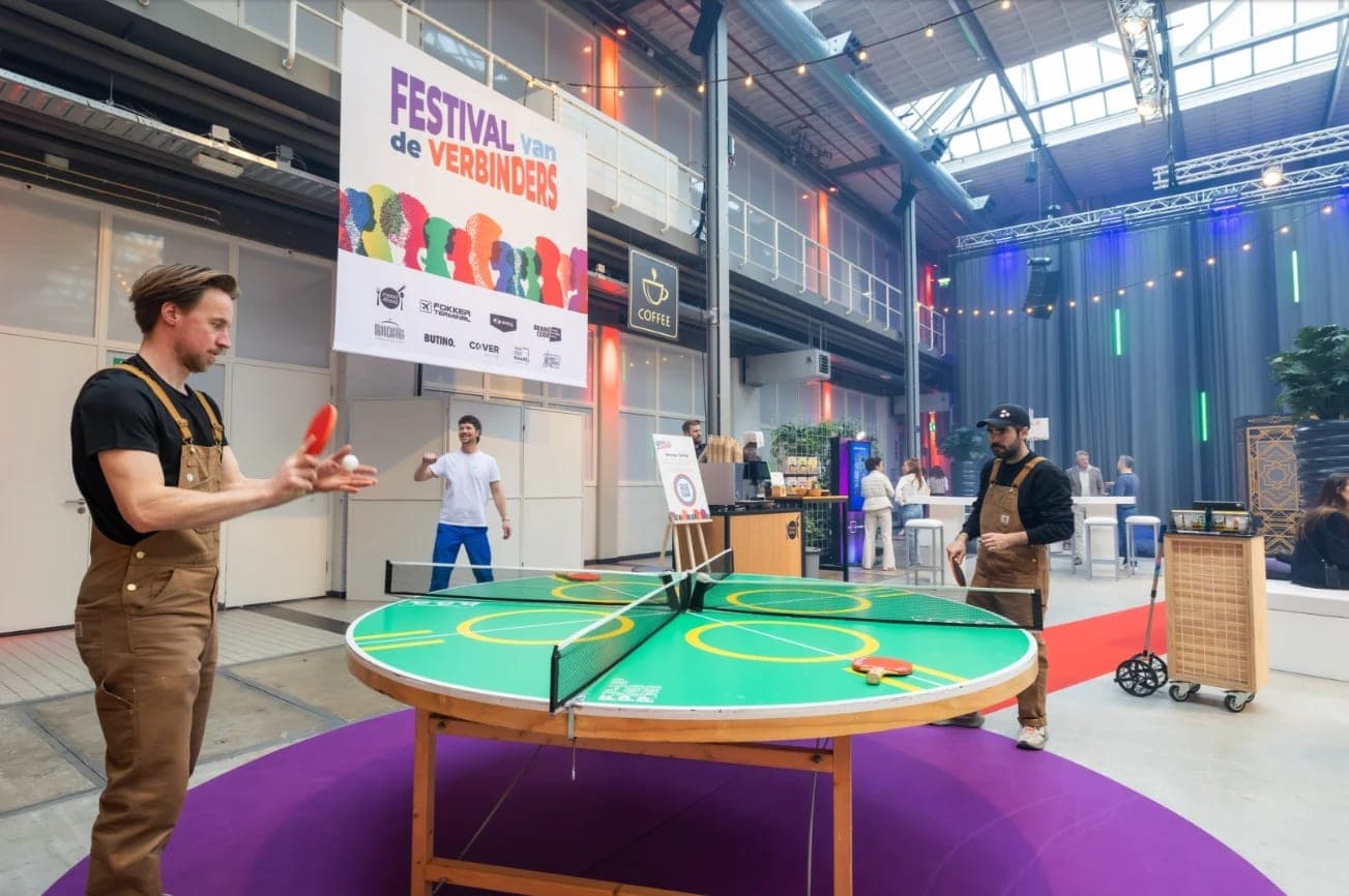 Bezoekers spelen tafeltennis aan een ronde tafel in de Fokker Terminal, met het spandoek van het Festival van de Verbinders op de achtergrond