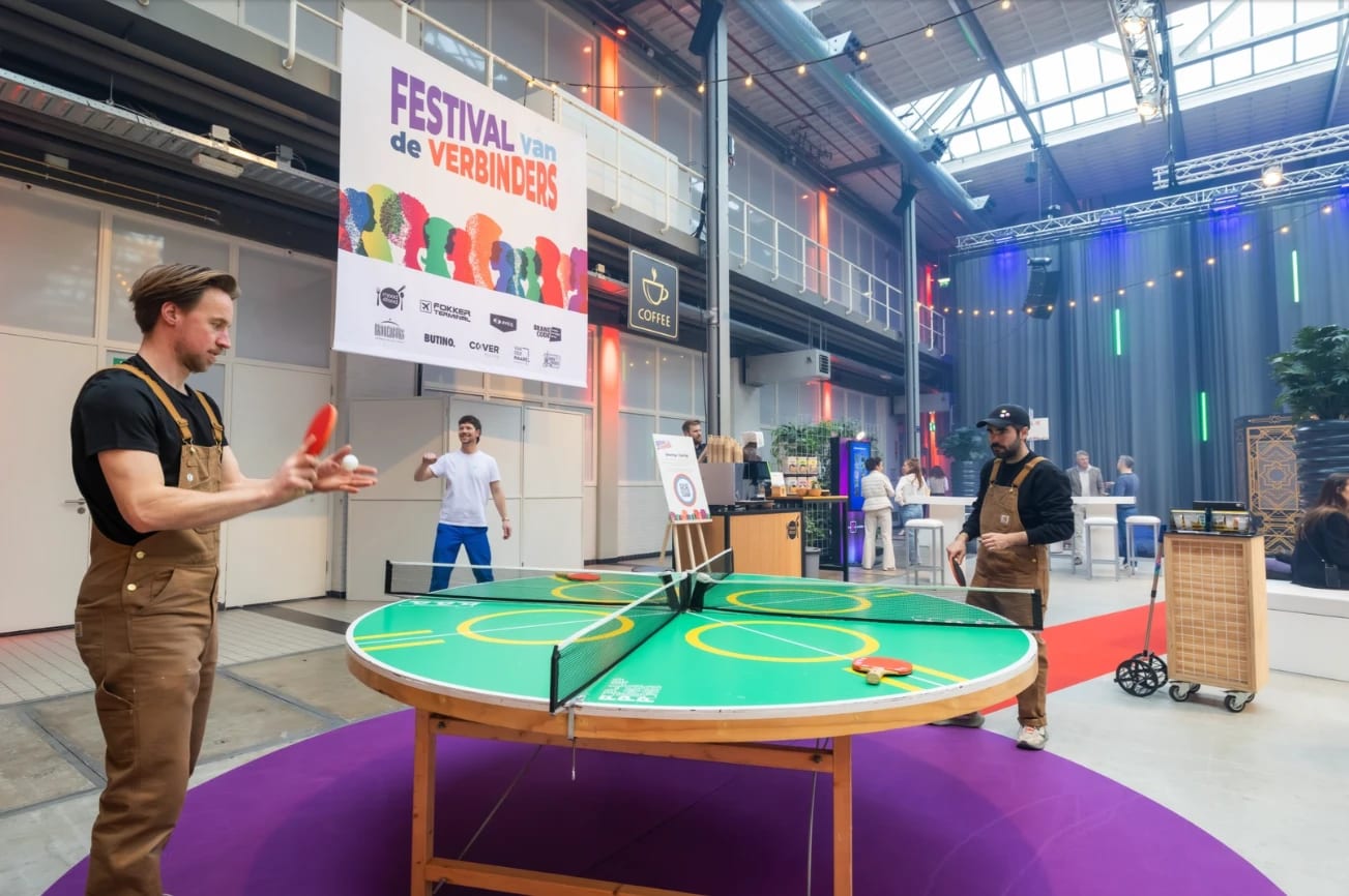 Bezoekers spelen tafeltennis aan een ronde tafel in de Fokker Terminal, met het spandoek van het Festival van de Verbinders op de achtergrond