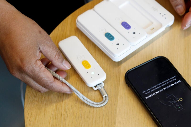 Hand legt een witte Speaksee-microfoon met gele knop op tafel naast een laadstation en een telefoon die vraagt de microfoon aan te zetten