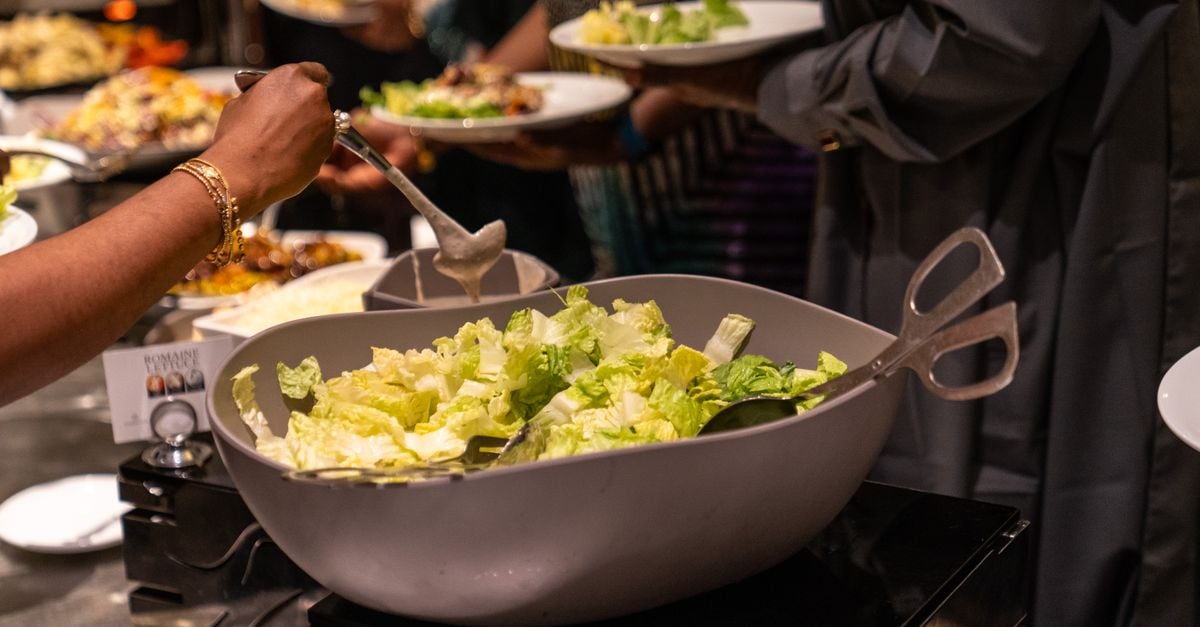 Een persoon serveert groene sla-salade op een buffet tijdens een sociale bijeenkomst binnenshuis.
