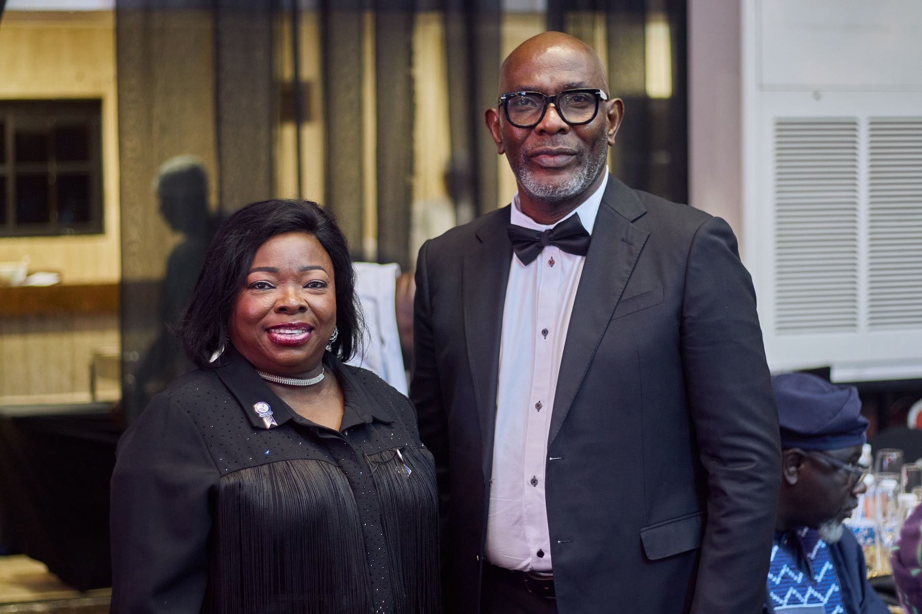 Two elegantly dressed individuals at a black tie event indoors, smiling for the camera.
