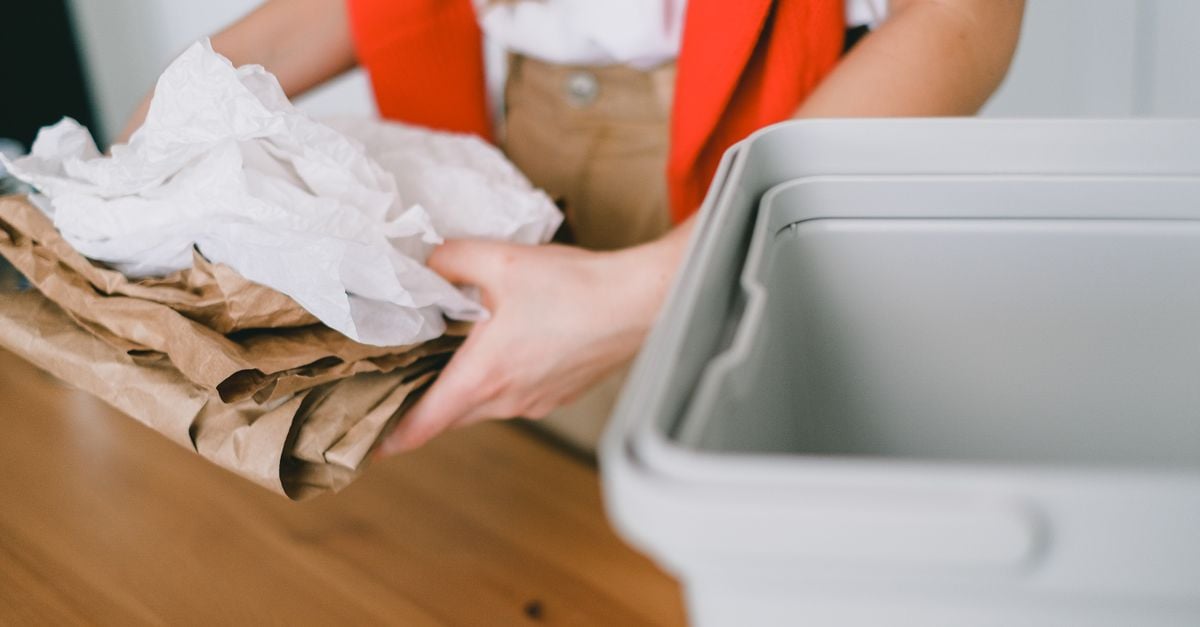 Crop anonymous female volunteer in casual clothes holding pile of paper bags while standing near wooden table with plastic container in light room