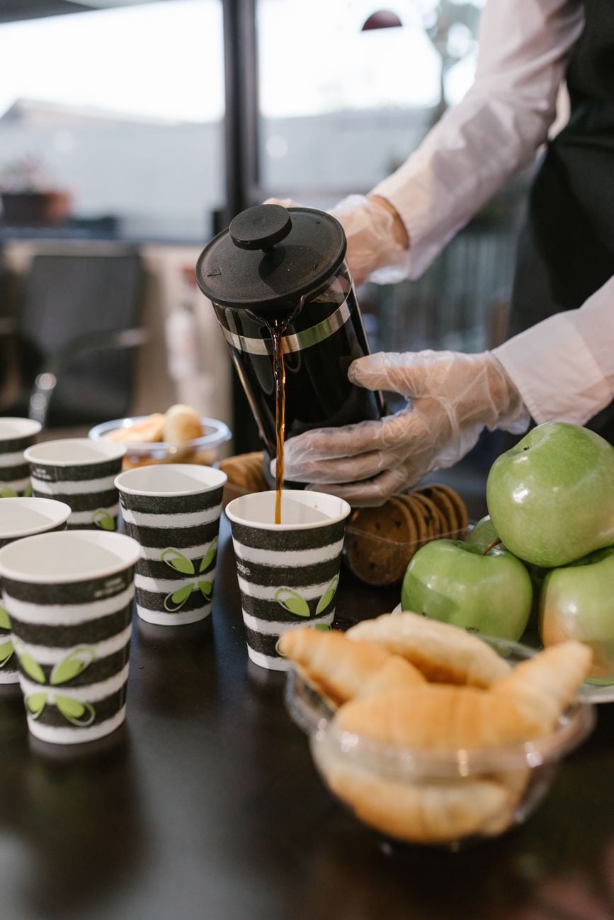 Barista giet koffie uit een French press in kopjes tijdens een ontbijt evenement met croissants en appels.
