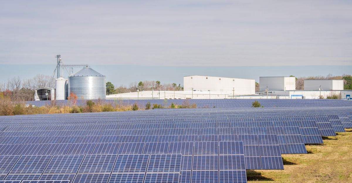 A large solar panel field with warehouses and silos in the background under a clear sky.