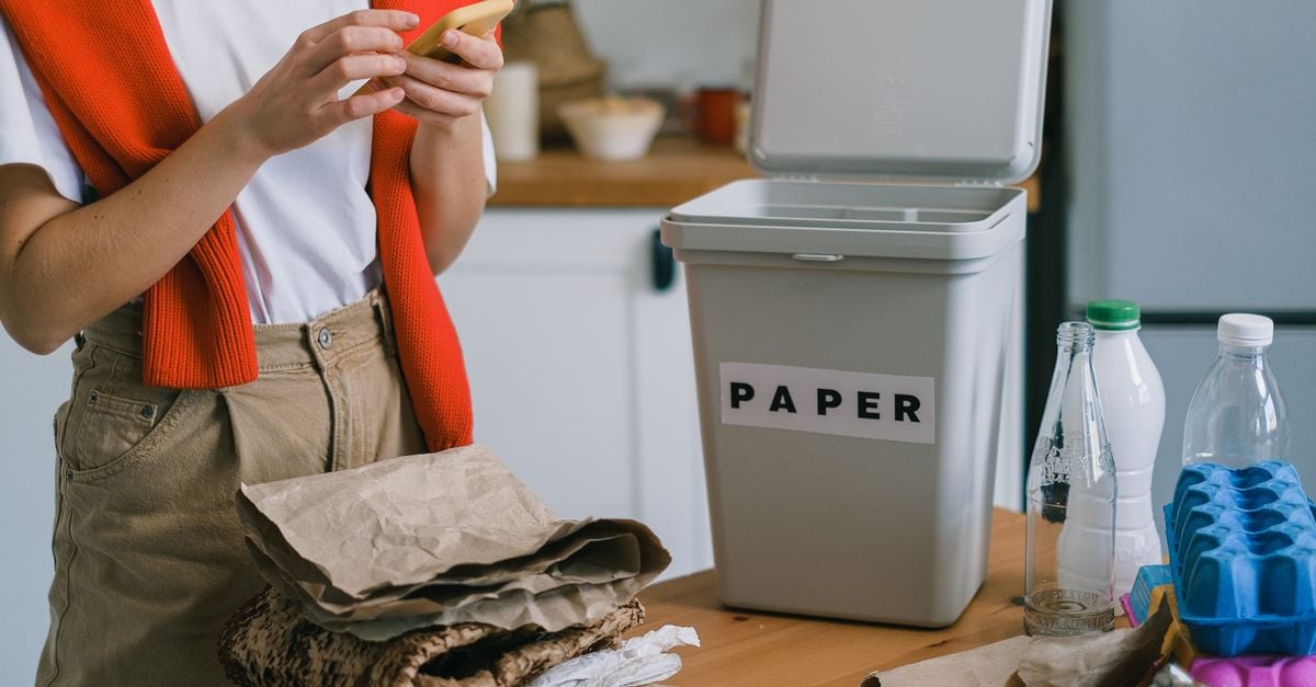 Crop unrecognizable person using smartphone while sorting waste on wooden table on blurred background