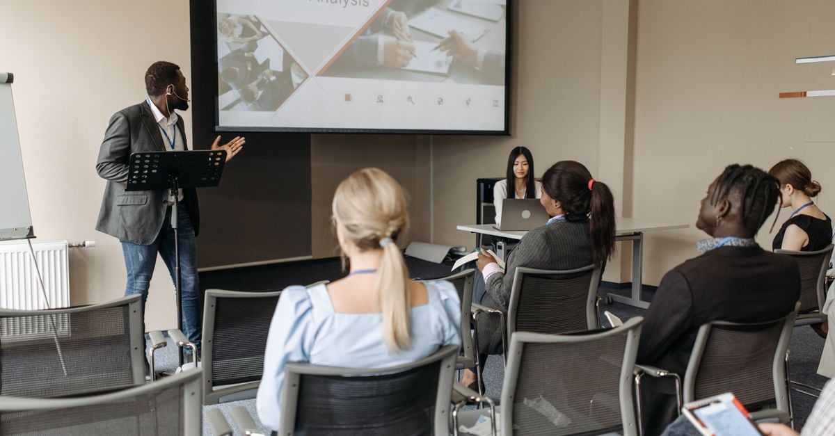 A diverse group attends a business analysis seminar with a presenter and projection screen in a modern conference room.