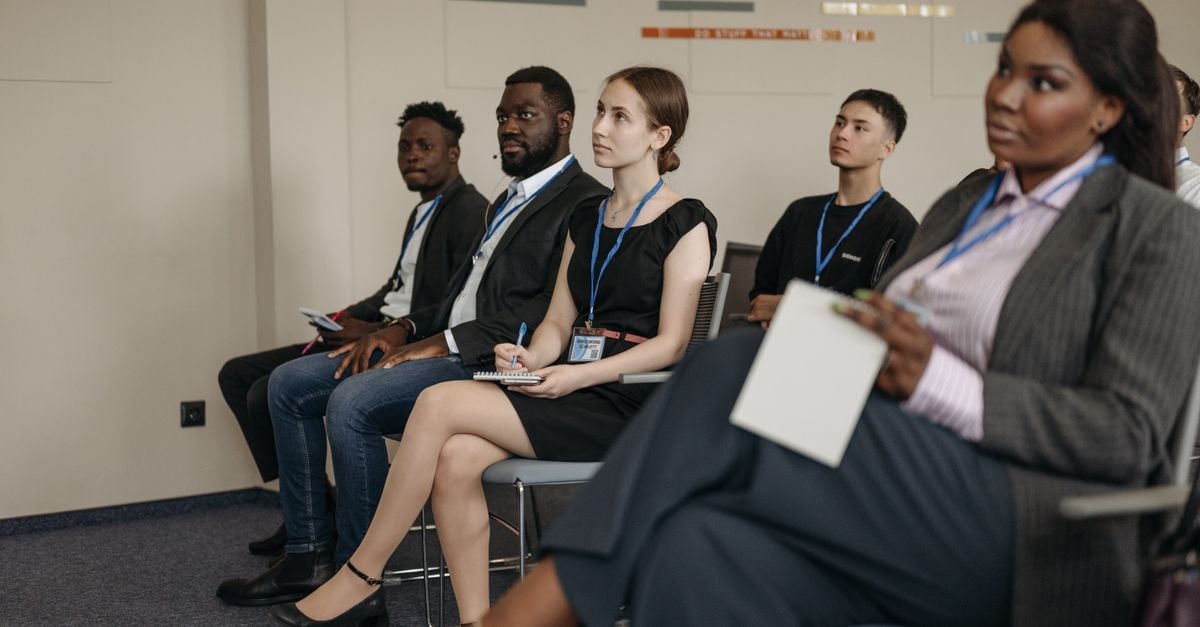 A diverse group of adults attentively listening in an indoor business meeting setting.
