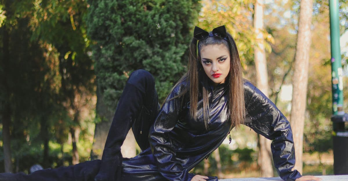Een vrouw in een zwart kostuum poseert zelfverzekerd buiten, omringd door herfstbomen.