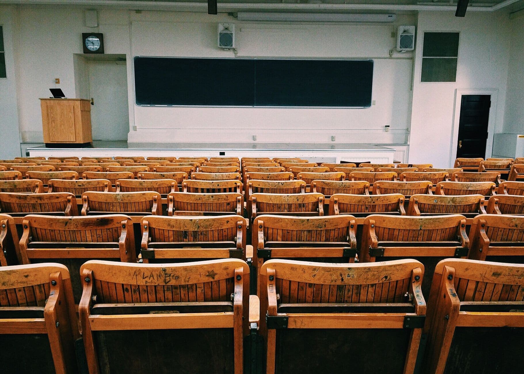 Een lege collegezaal met houten stoelen en een groot prikbord.