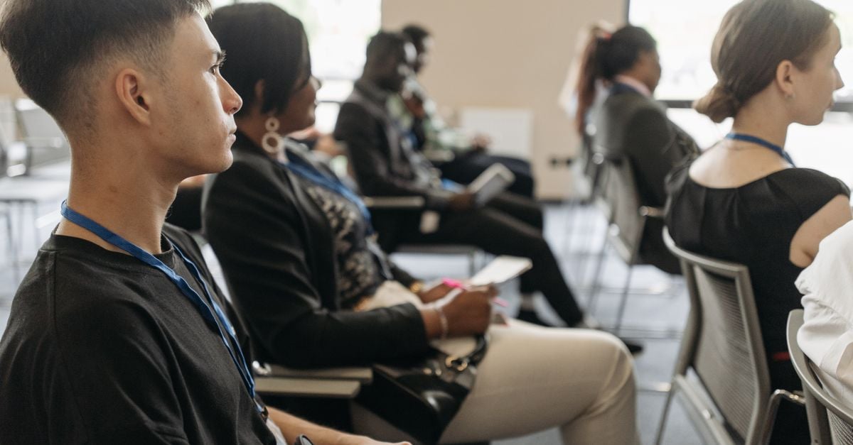 A diverse group of adults attentively listening at a conference setting, featuring name tags and formal attire.