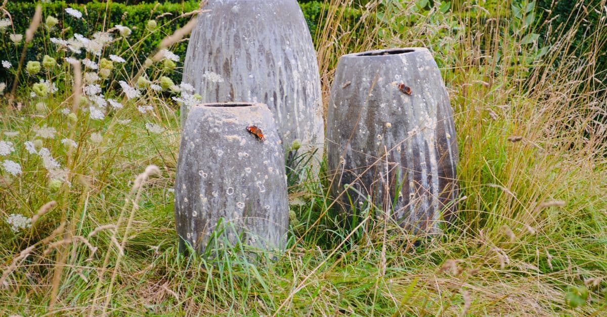 Drie grote stenen plantenbakken, omgeven door wilde grassen en witte bloemen, in een buitentuin.