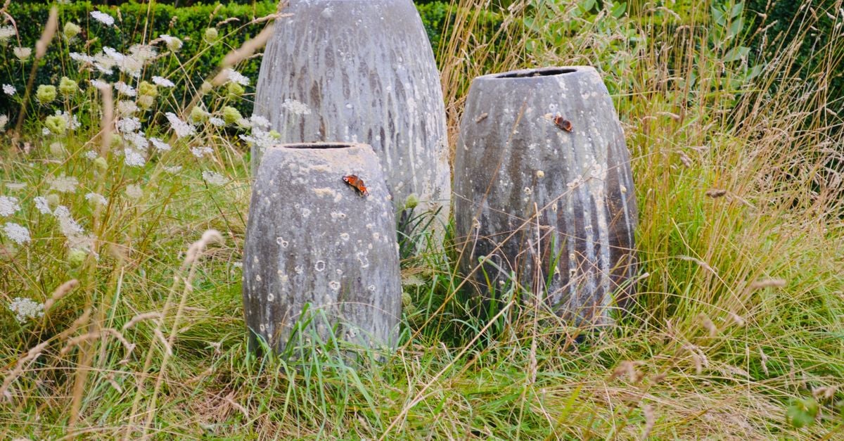 Drie grote stenen plantenbakken, omgeven door wilde grassen en witte bloemen, in een buitentuin.