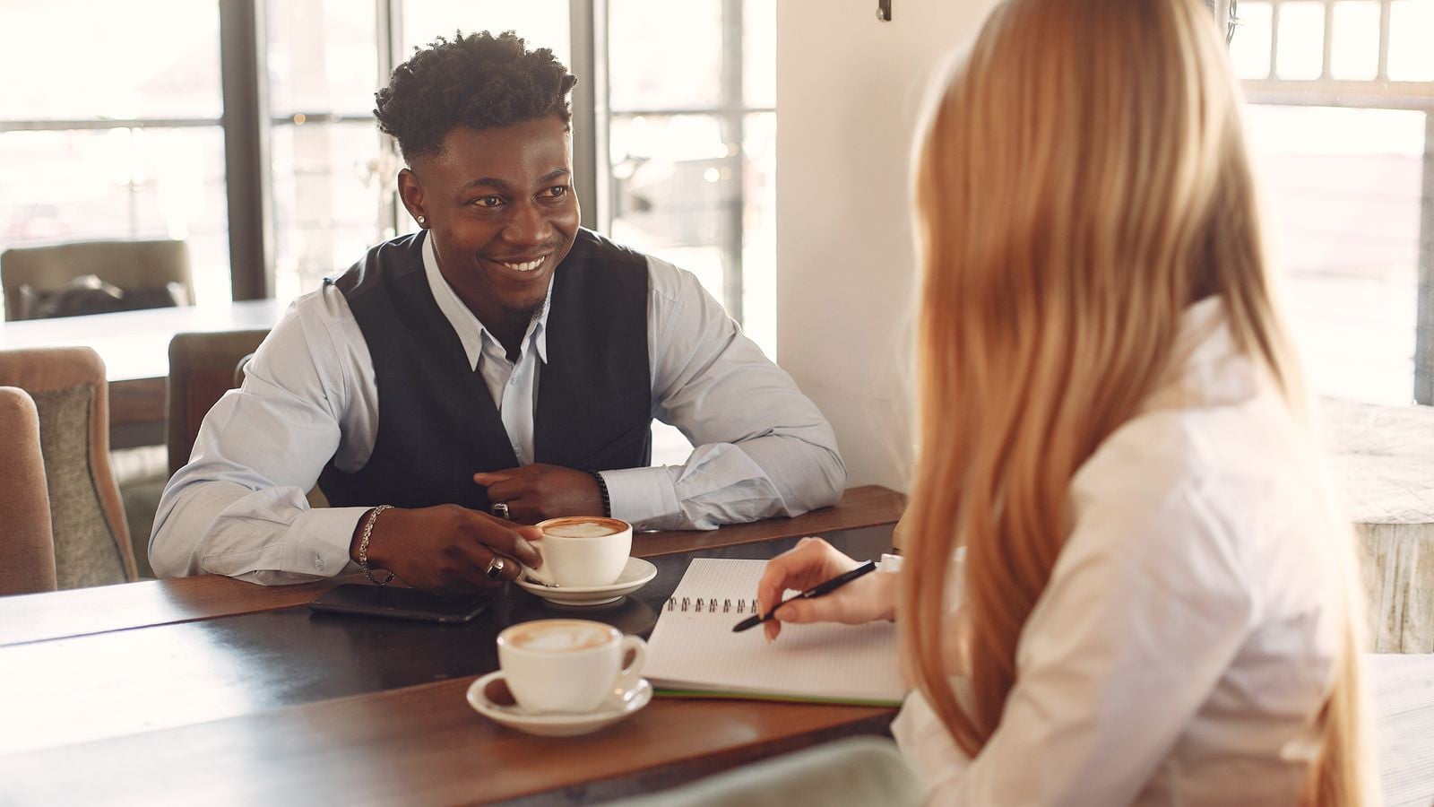 Twee mensen in gesprek bij een kop koffie — de een praat, de ander luistert en maakt aantekeningen